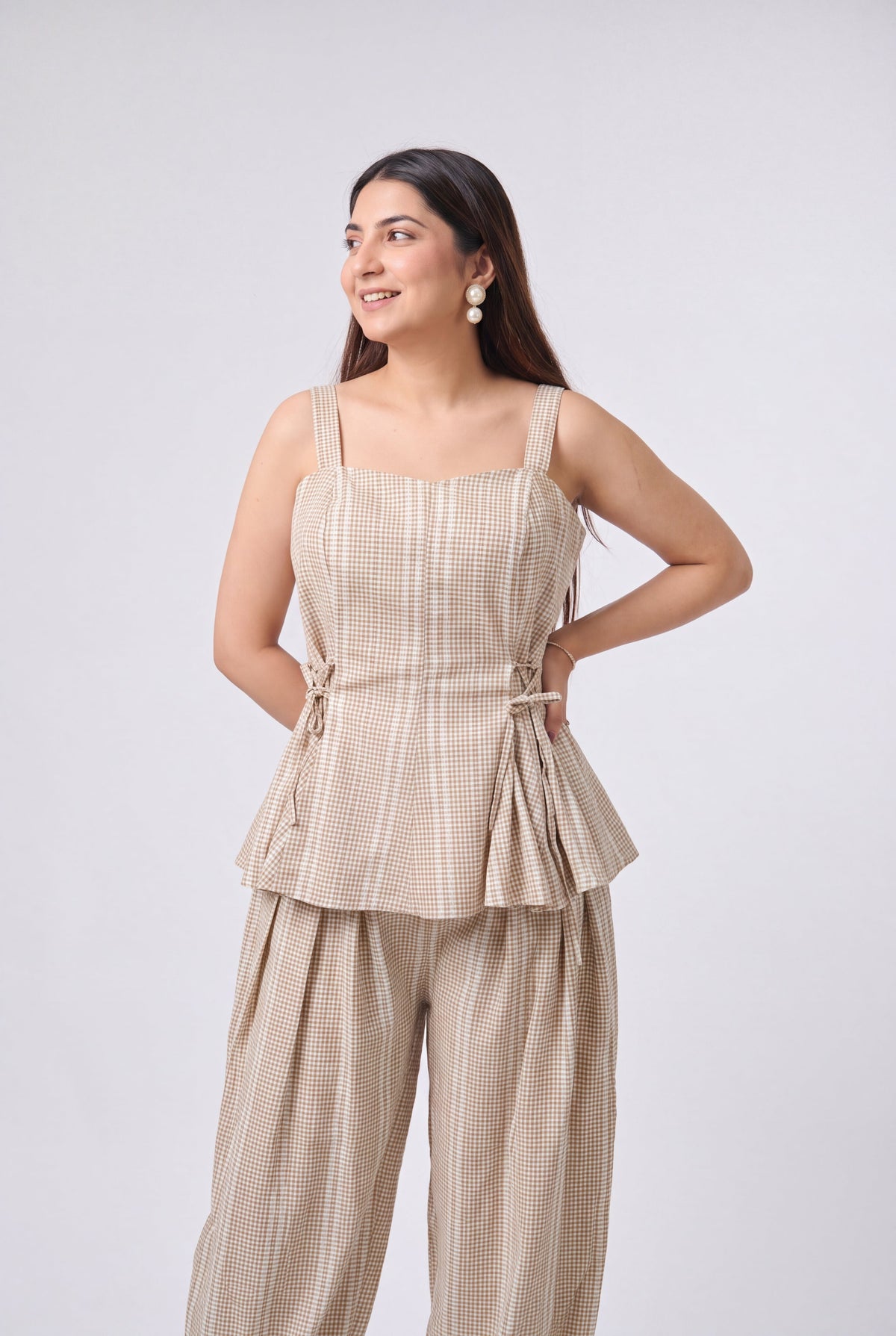 A woman modeling a beige and white checkered two-piece set consisting of a sleeveless peplum top with side ties and matching wide-leg pants, photographed against a light gray studio background.