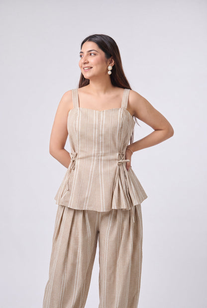 A woman modeling a beige and white checkered two-piece set consisting of a sleeveless peplum top with side ties and matching wide-leg pants, photographed against a light gray studio background.