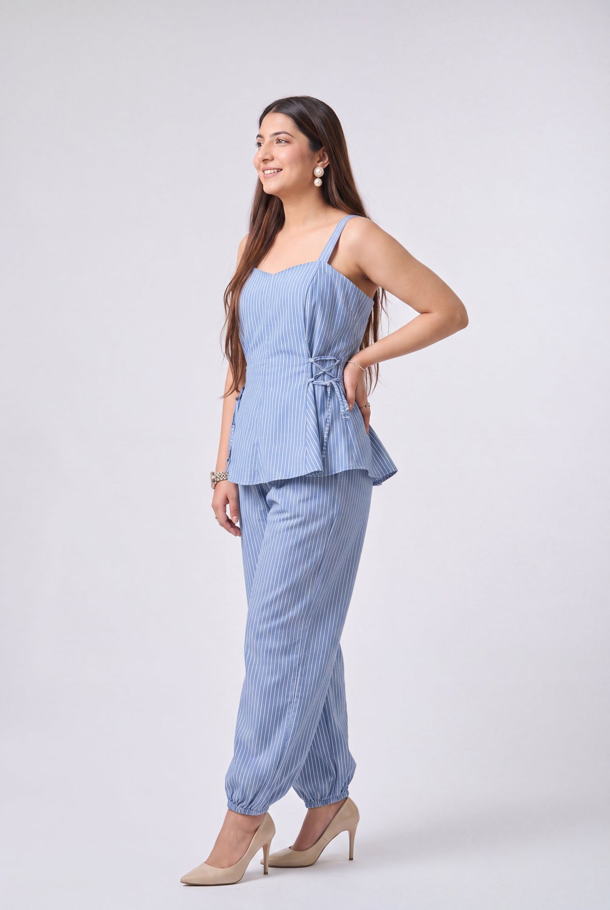 A woman modeling a light blue and white vertically striped two-piece set consisting of a sleeveless peplum top with a tie detail at the waist and matching cropped pants, paired with beige high heels against a light gray studio background.