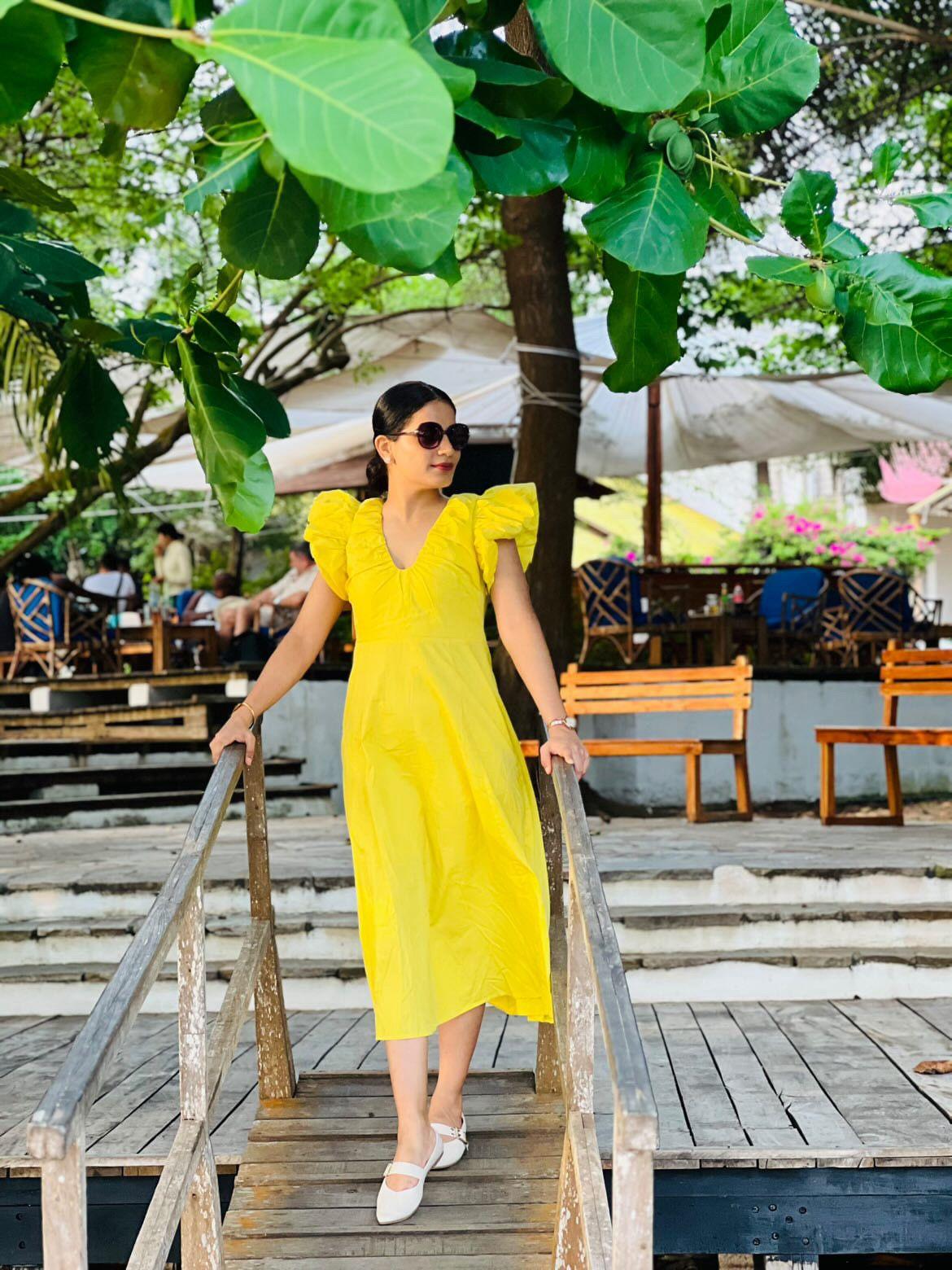 Woman in a yellow dress standing on a wooden platform with greenery and people in the background