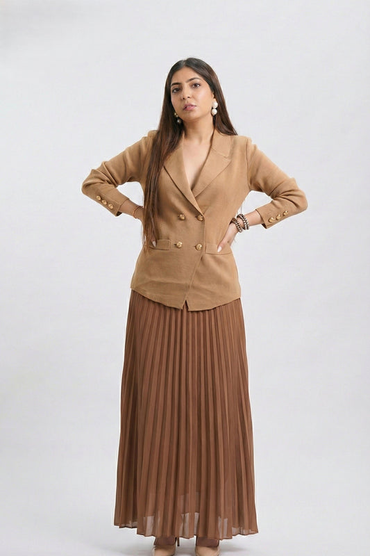 A woman modeling a tan double-breasted blazer paired with a brown pleated maxi skirt, standing against a light gray studio background.