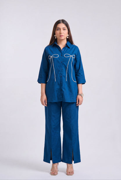 A woman modeling a matching blue two-piece set consisting of a long-sleeve button-up shirt with white bow embroidery and matching wide-leg trousers, shot against a light gray studio background.
