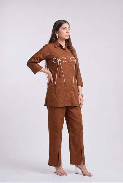 A woman modeling a brown two-piece linen pajama set consisting of a long-sleeve button-up top with white cord details and matching wide-leg pants, shot against a light gray studio background.