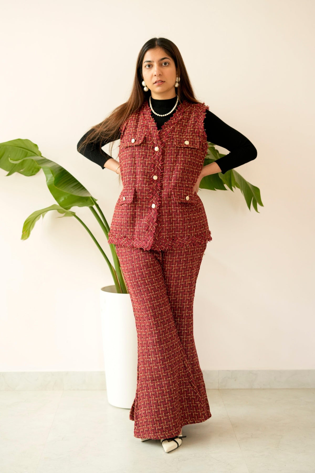 Woman wearing a red checkered outfit with a vest and pants, standing against a white wall with a plant.