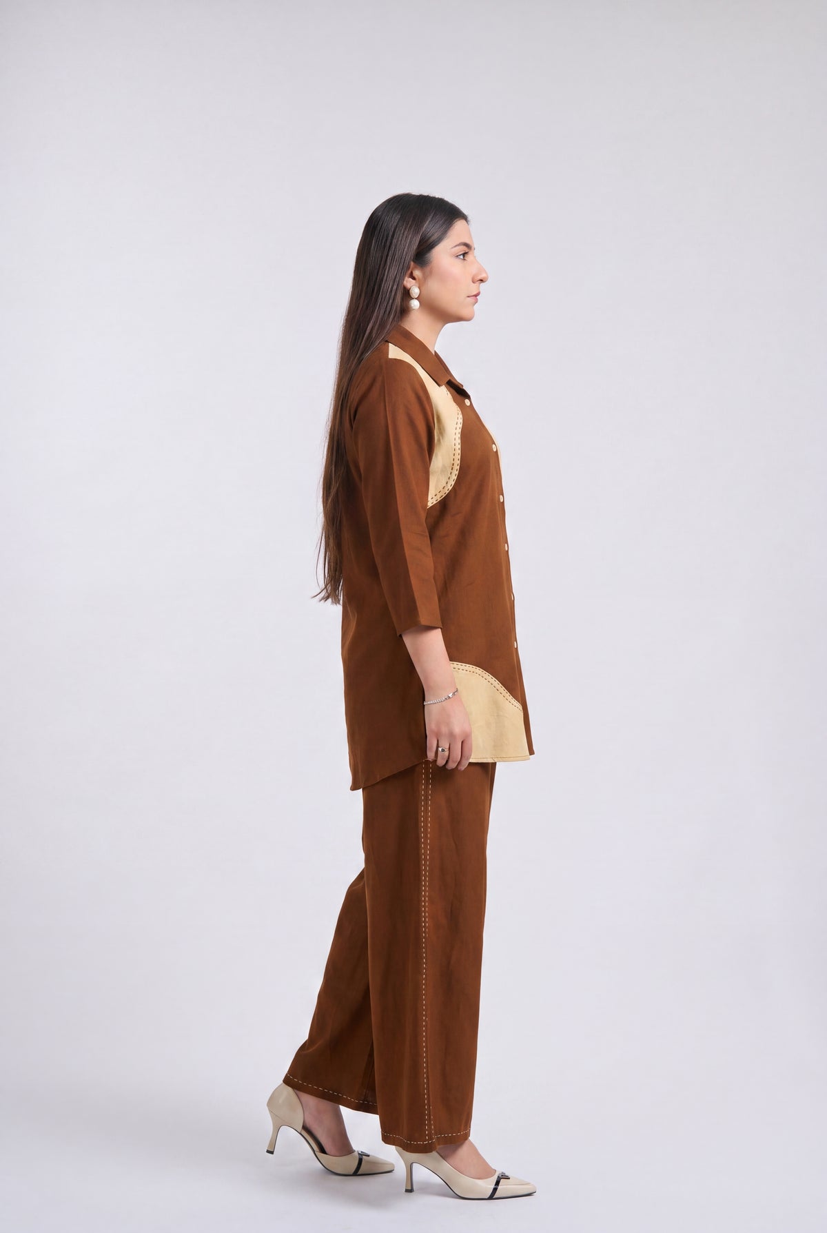 A woman stands in profile wearing a brown two-piece outfit with contrasting beige panels, paired with beige and white pointed-toe heels, against a light gray studio background.