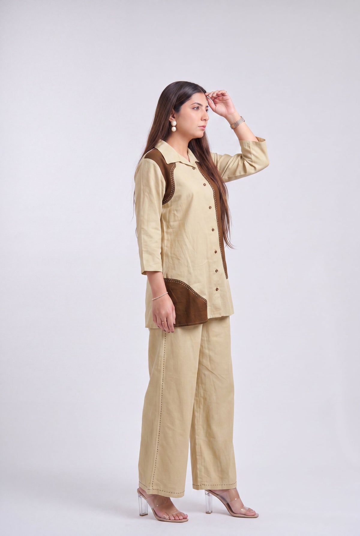 A woman modeling a beige two-piece linen set consisting of a long-sleeve button-up shirt with brown contrast panels and matching wide-leg trousers, set against a plain light gray studio background.