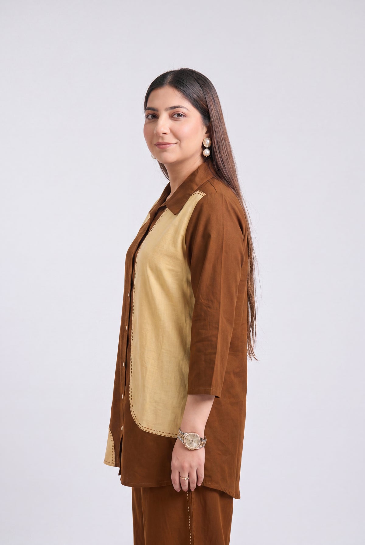 A woman modeling a brown and beige kurta set against a plain light gray studio background. She is shown in profile, wearing a brown button-up jacket over a beige tunic with matching brown pants.