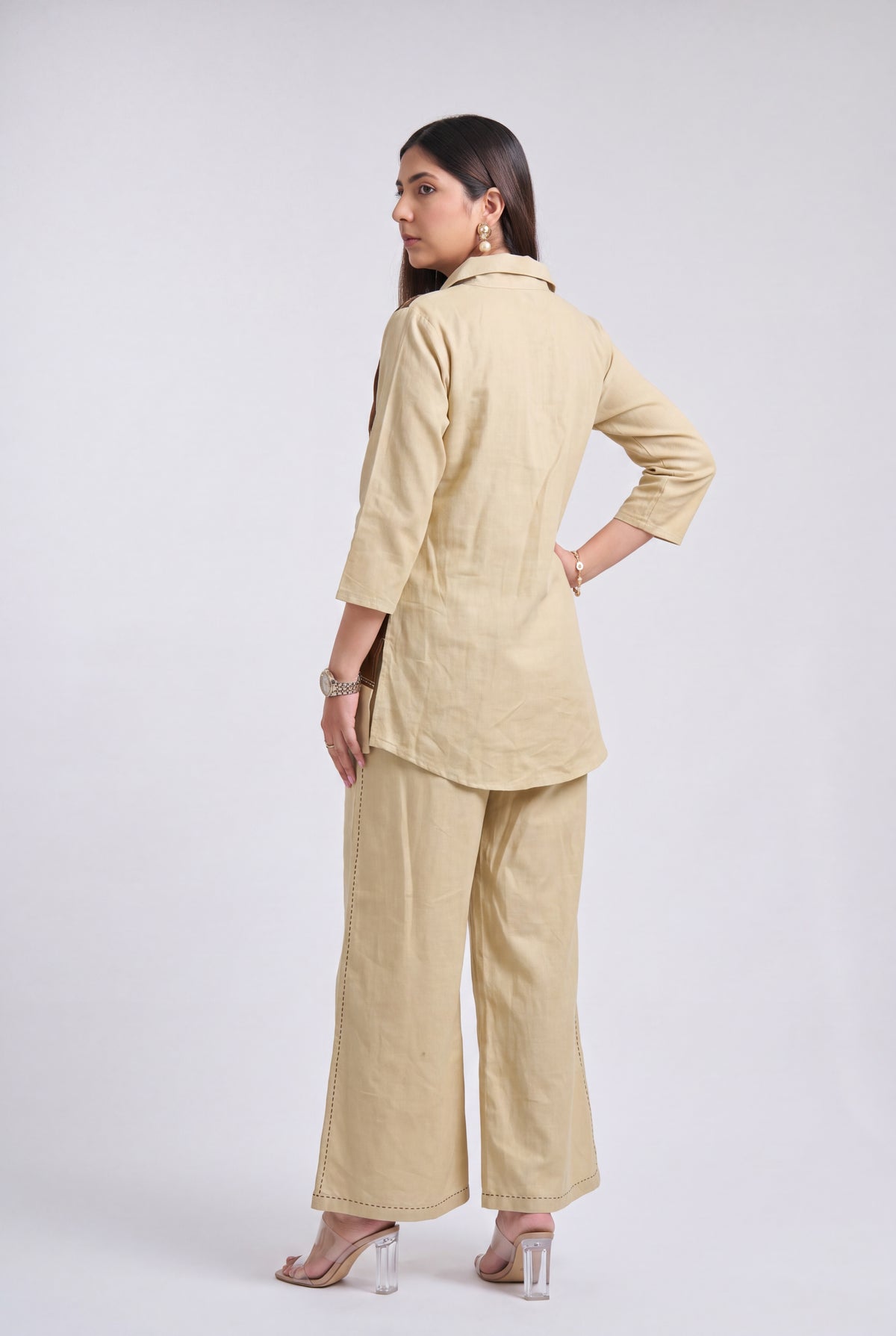 A woman modeling a light beige linen two-piece set consisting of a long-sleeve collared tunic top and matching wide-leg trousers, shown from a rear three-quarter angle against a light gray studio background.