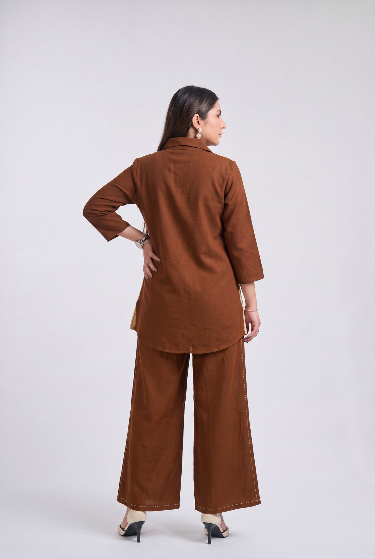 A woman is shown from the back wearing a matching brown two-piece outfit consisting of a long-sleeve top and wide-leg trousers, paired with black heels, against a light gray studio background.