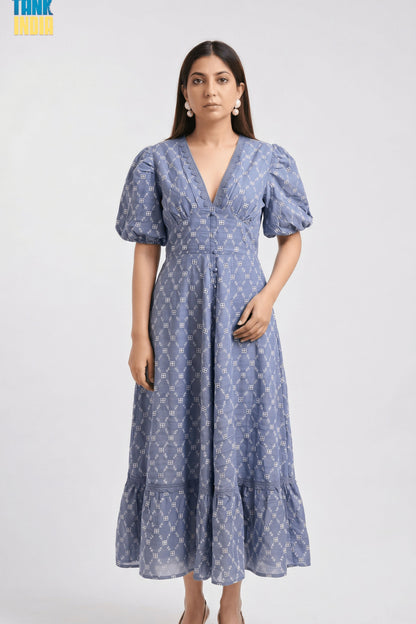 A woman modeling a blue midi dress with a white geometric pattern, puff sleeves, and a V-neckline against a light gray studio background.