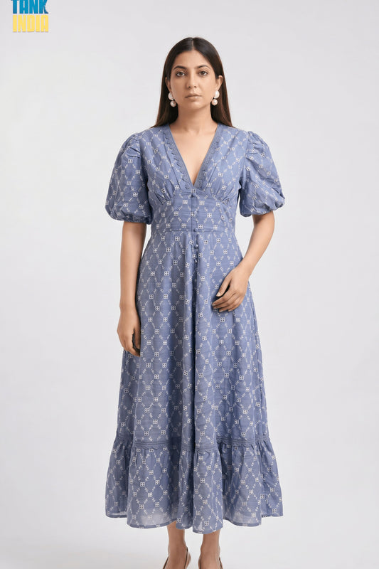 A woman modeling a blue midi dress with a white geometric pattern, puff sleeves, and a V-neckline against a light gray studio background.