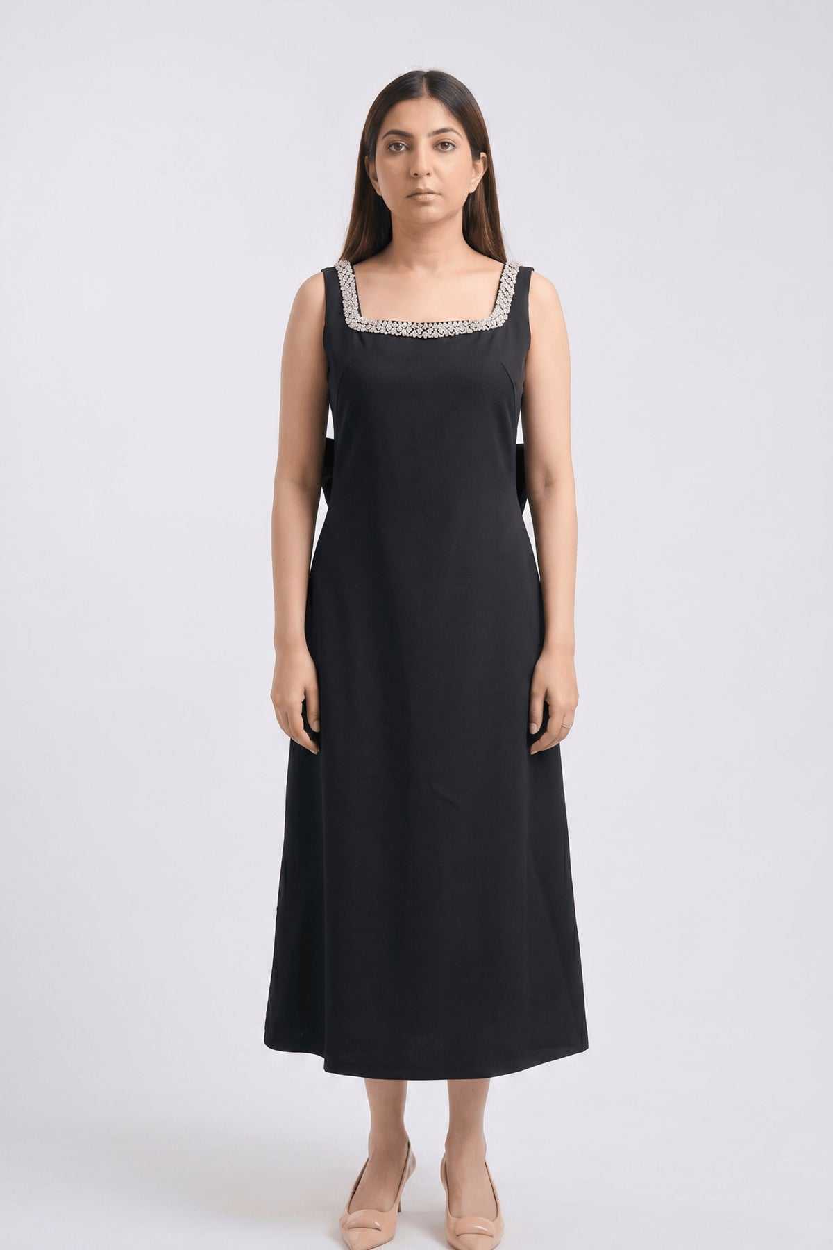 A woman modeling a sleeveless black midi dress with a square neckline decorated with silver and white beaded trim, standing against a light gray studio background.