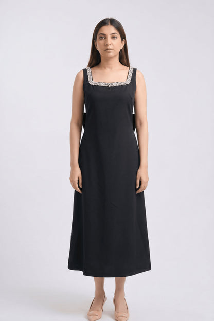 A woman modeling a sleeveless black midi dress with a square neckline decorated with silver and white beaded trim, standing against a light gray studio background.
