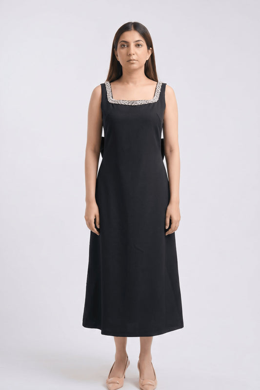 A woman modeling a sleeveless black midi dress with a square neckline decorated with silver and white beaded trim, standing against a light gray studio background.
