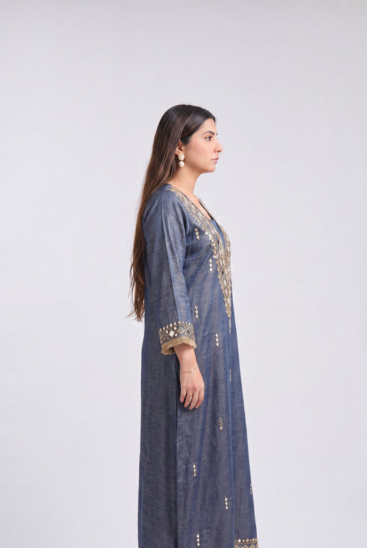 A woman stands in profile wearing a long blue-gray kurta with gold embroidery and mirror work on the neckline, cuffs, and hem, paired with white heeled sandals against a light gray studio background.