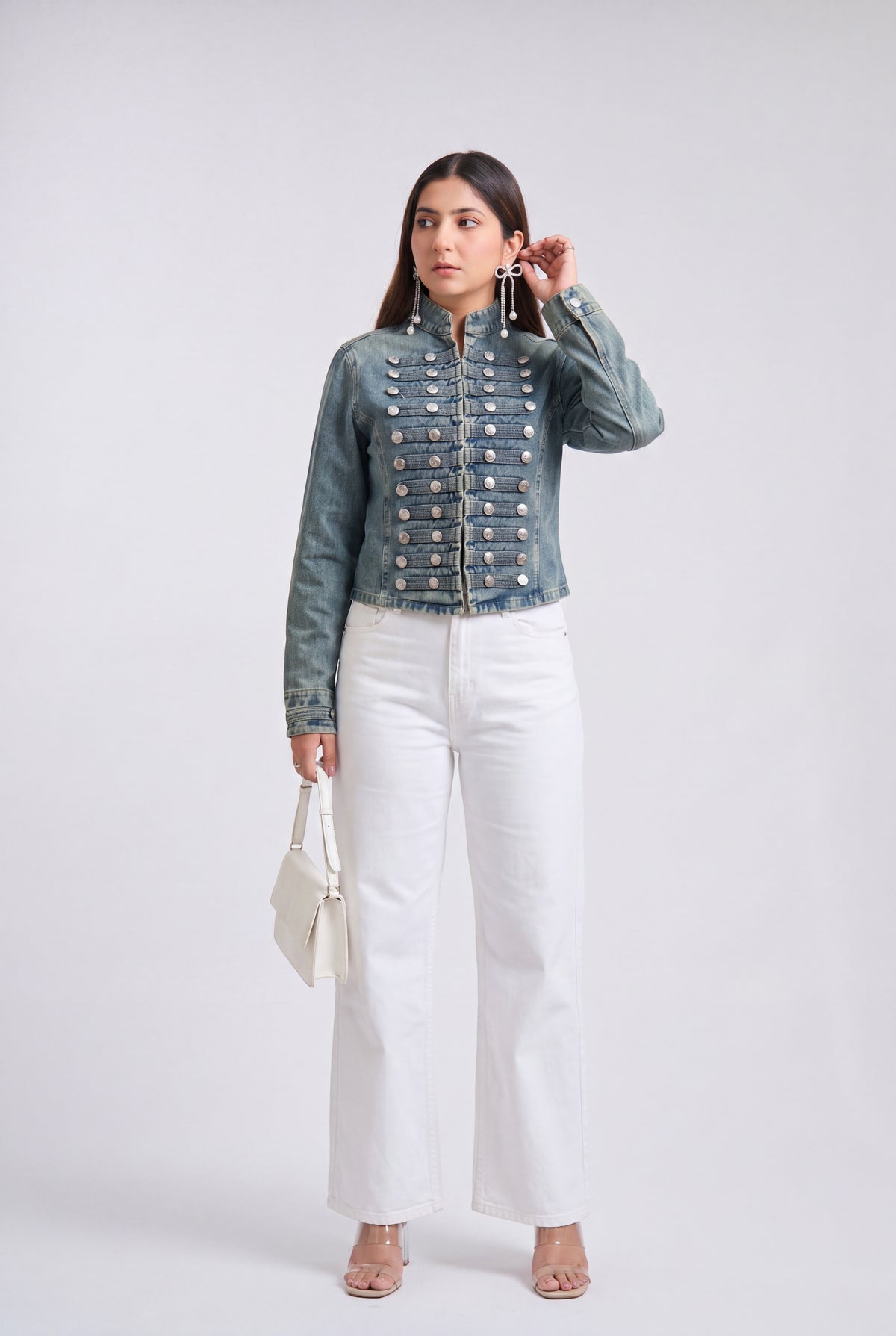 A woman modeling an denim blue military-style jacket with silver buttons, paired with black wide-leg pants and clear high-heeled sandals, against a light gray studio background.