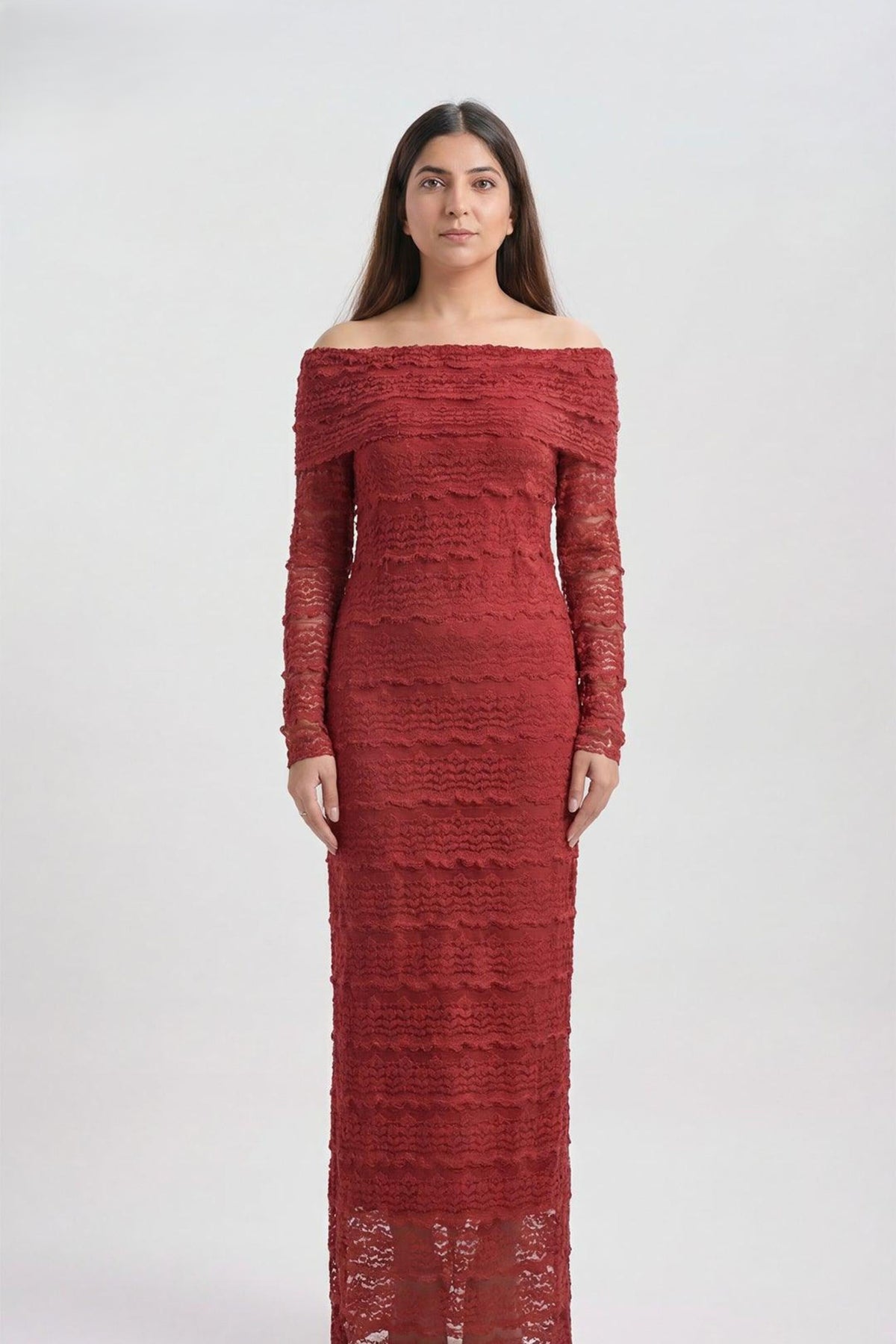 A woman modeling a long-sleeve off-the-shoulder maxi dress in textured red lace against a plain light gray studio background.