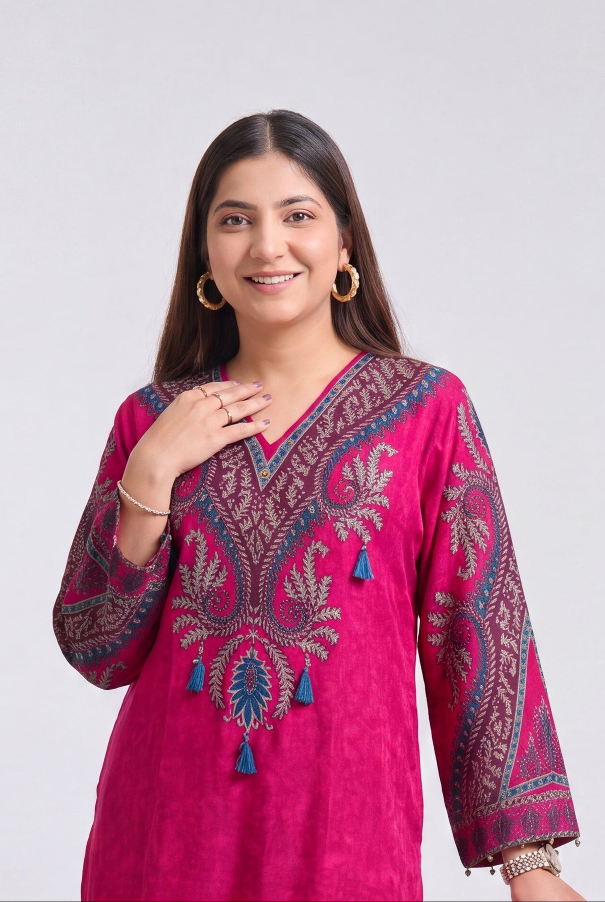 A woman modeling a bright pink traditional kurta set with detailed embroidery, standing next to a wooden stool against a light gray background.