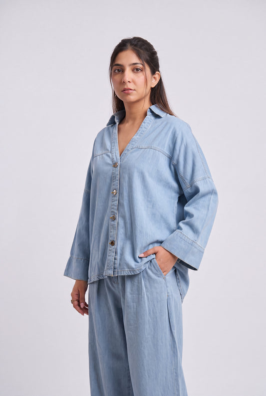 A woman modeling a light blue denim button-up shirt and matching wide-leg pants against a plain light gray studio background.