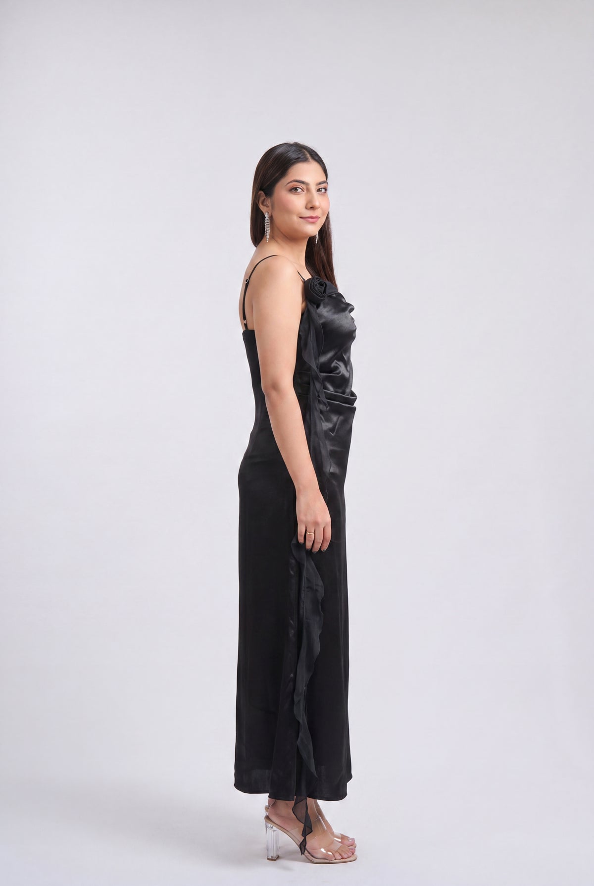 A woman modeling a black satin-like sleeveless maxi dress with a draped, gathered bodice and side slit, standing in clear high-heeled sandals against a light gray studio background.