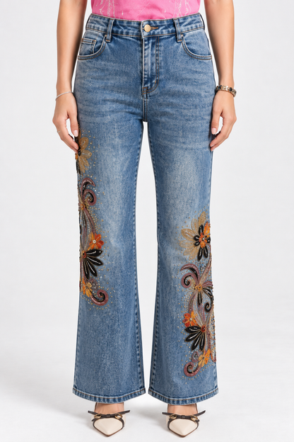 A model wearing high-waisted, wide-leg blue denim jeans with colorful floral embroidery on the front of each leg, paired with a pink top and beige strappy heels against a white studio background.