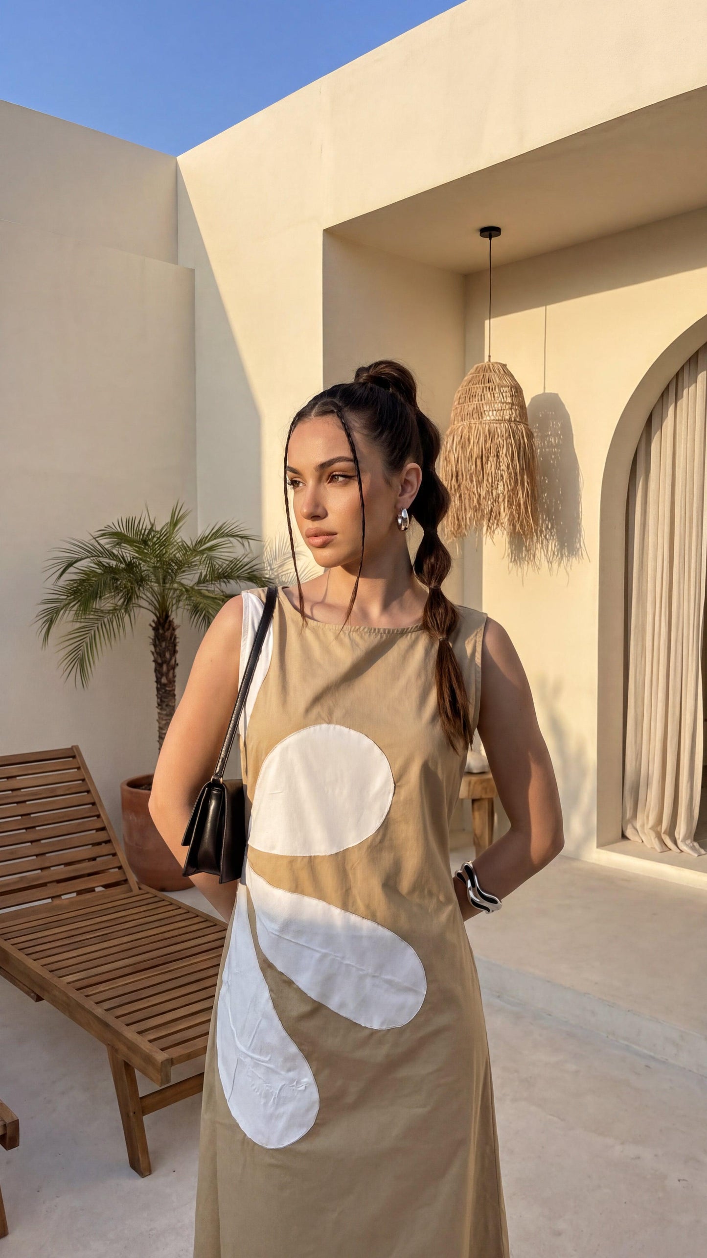Woman in a beige dress with white patterns standing in a sunlit outdoor setting.