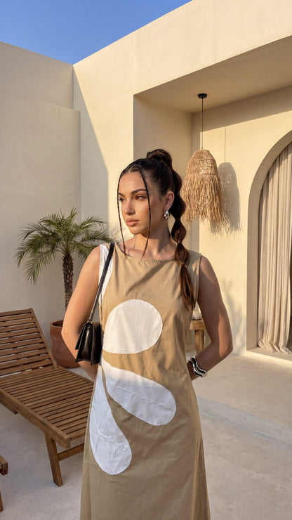 Woman in a beige dress with white patterns standing in a sunlit outdoor setting.