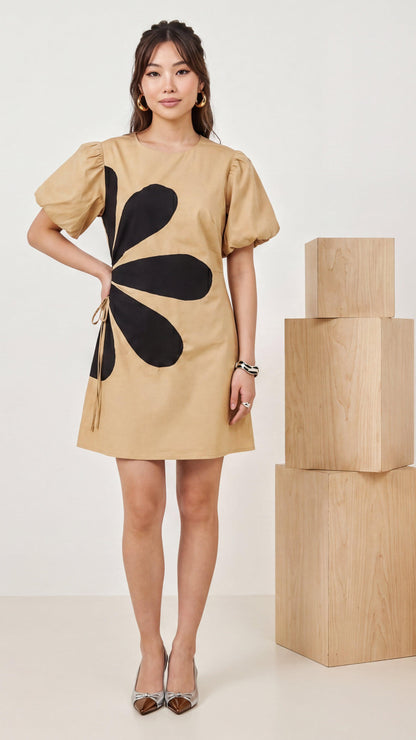 Woman wearing a beige dress with a large black flower design, standing next to stacked wooden boxes.