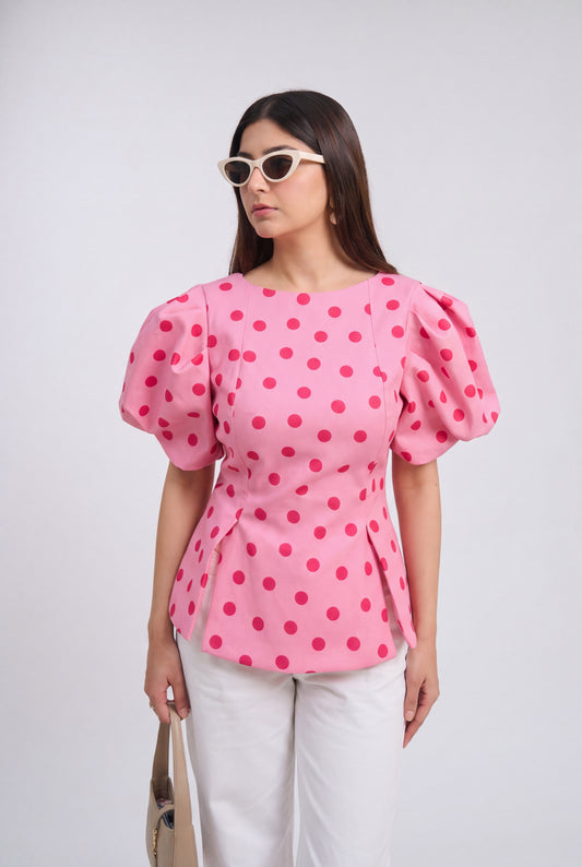 A woman modeling a pink puff-sleeve blouse with a bold magenta polka dot pattern, paired with white pants and white-framed sunglasses, against a light gray studio background.