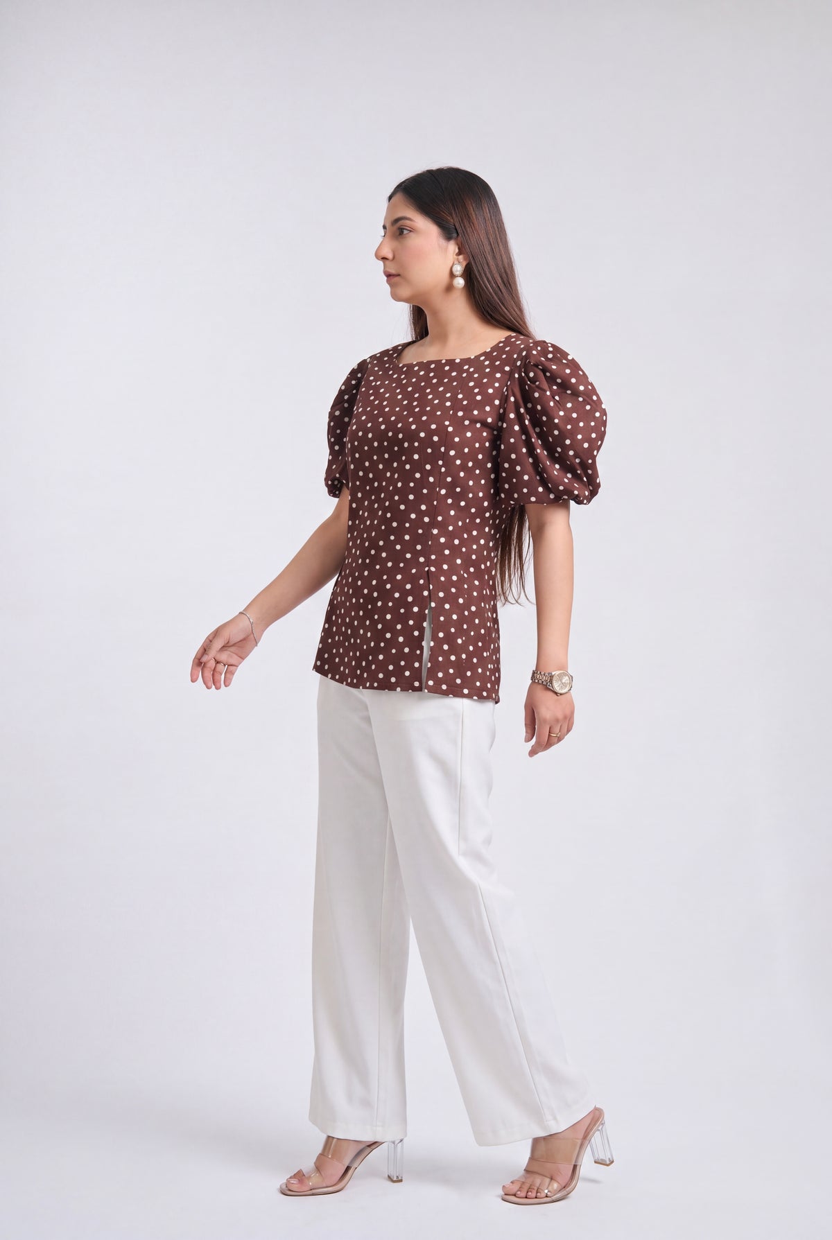 A woman modeling a brown puff-sleeve top with a white polka dot pattern, paired with white wide-leg pants and clear high-heeled sandals, set against a plain light gray studio background.