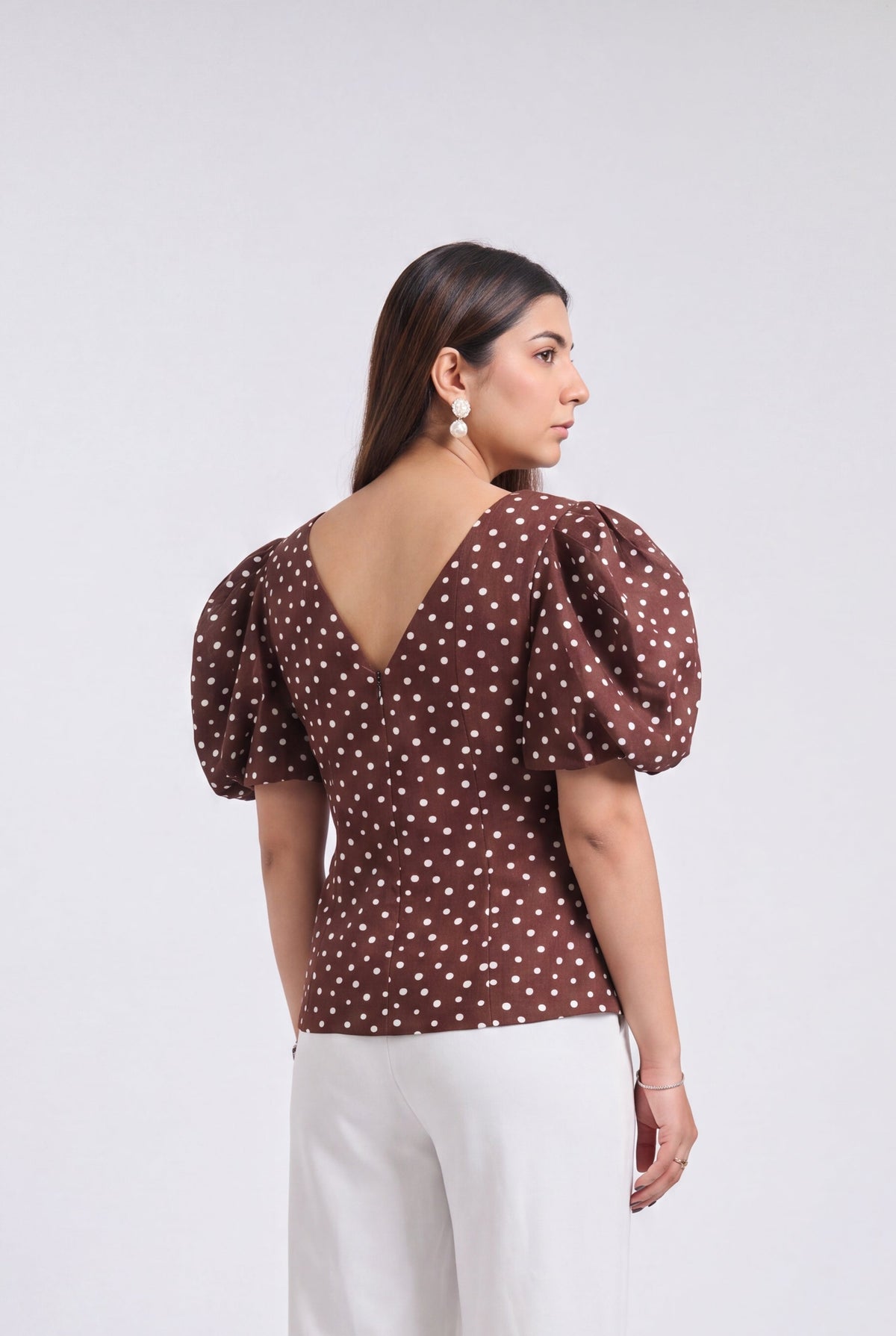 A woman is shown from the back wearing a brown puff-sleeve top with a white polka dot pattern and white wide-leg trousers, standing against a light gray studio background.
