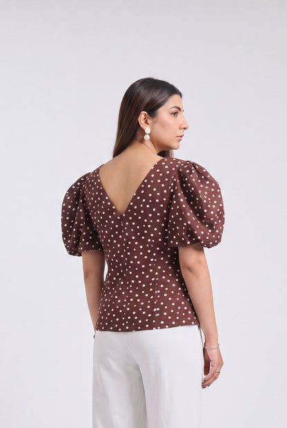 A woman is shown from the back wearing a brown puff-sleeve top with a white polka dot pattern and white wide-leg trousers, standing against a light gray studio background.