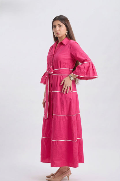 A woman modeling a bright pink tiered maxi shirt dress with white piping details, a matching waist tie belt, and flared sleeves, set against a plain light gray studio background.