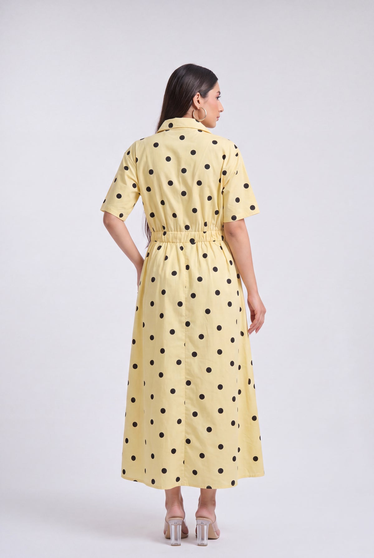 A woman viewed from the back wearing a pale yellow short-sleeve midi dress with black polka dots and a matching tie belt, photographed against a light gray studio background.