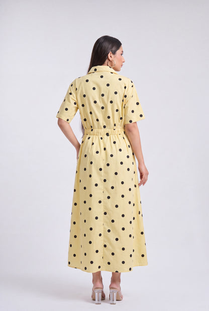 A woman viewed from the back wearing a pale yellow short-sleeve midi dress with black polka dots and a matching tie belt, photographed against a light gray studio background.