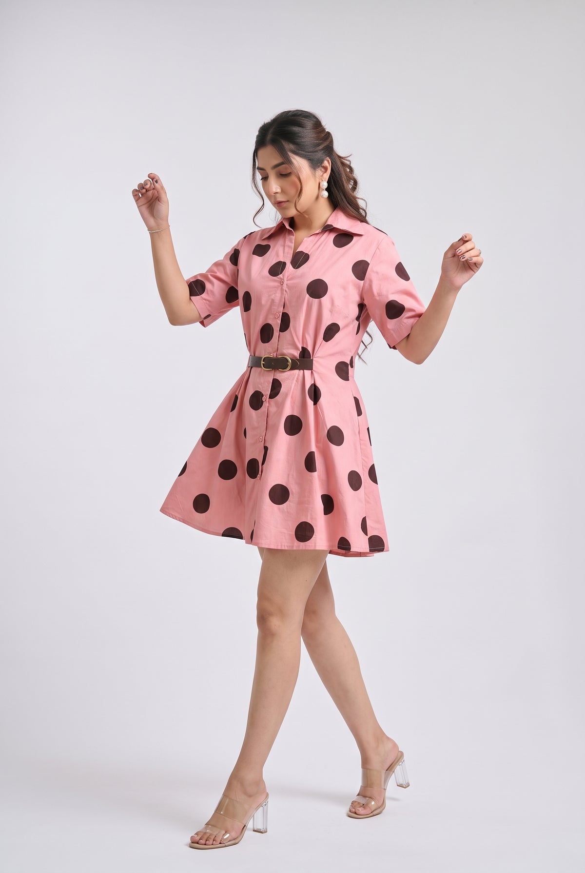 A woman modeling a pink short-sleeve mini dress with large black polka dots and a thin dark belt, posed against a light gray studio background.