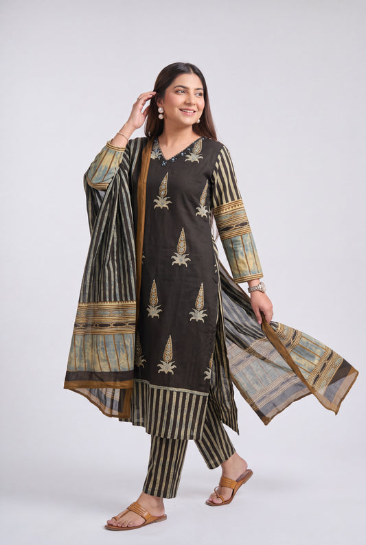A woman modeling a traditional three-piece salwar kameez suit in dark brown and beige. The kurta features vertical beige stripes and embroidered motifs, paired with matching striped trousers and a sheer dupatta with beige and gold patterns, set against a plain light gray studio background.