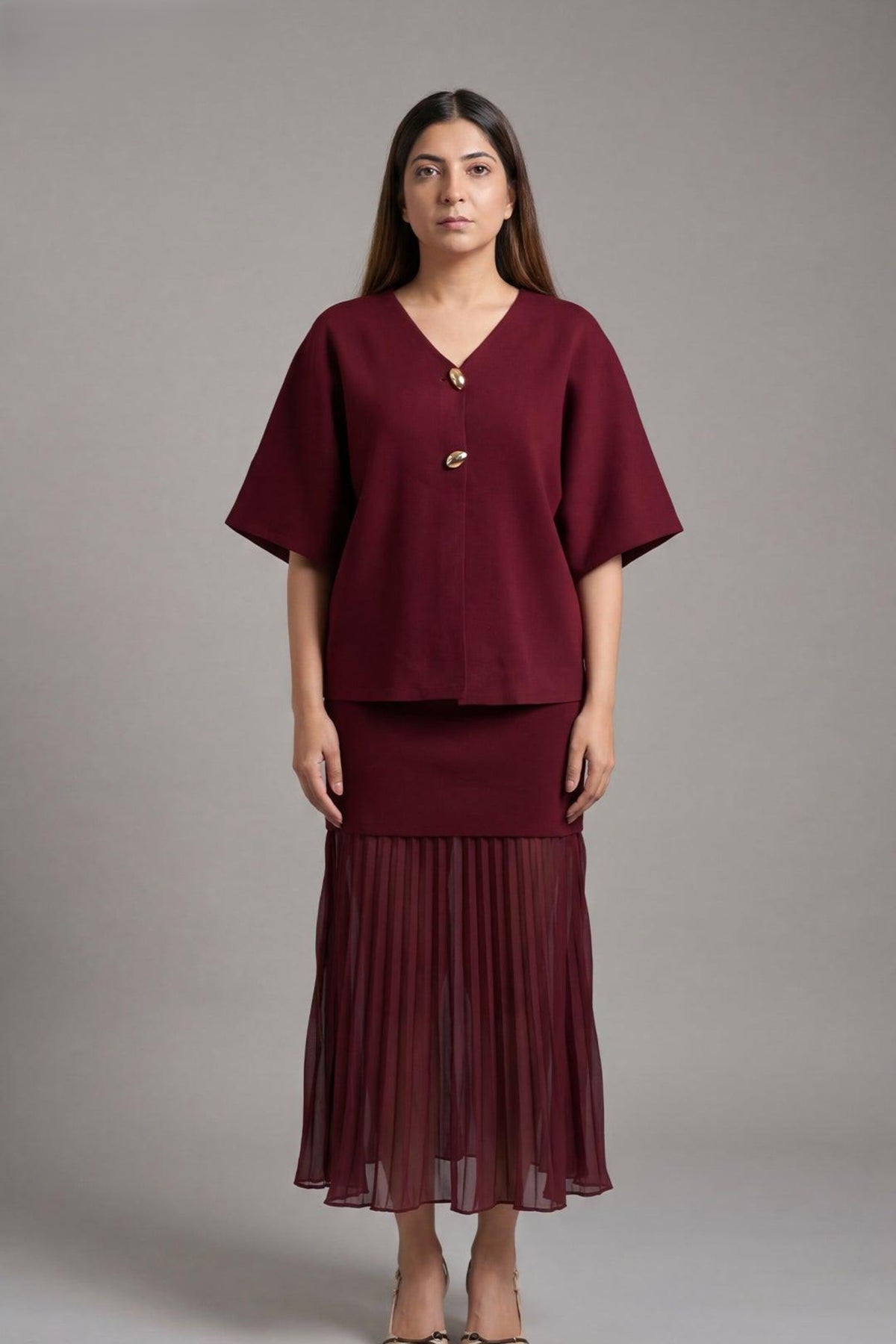 A woman modeling a matching burgundy two-piece set consisting of a short-sleeve V-neck top with a decorative front button and a pleated midi skirt, shot against a solid gray studio background.