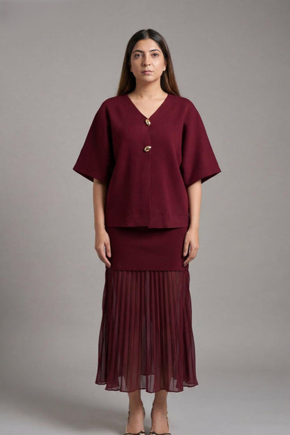 A woman modeling a matching burgundy two-piece set consisting of a short-sleeve V-neck top with a decorative front button and a pleated midi skirt, shot against a solid gray studio background.