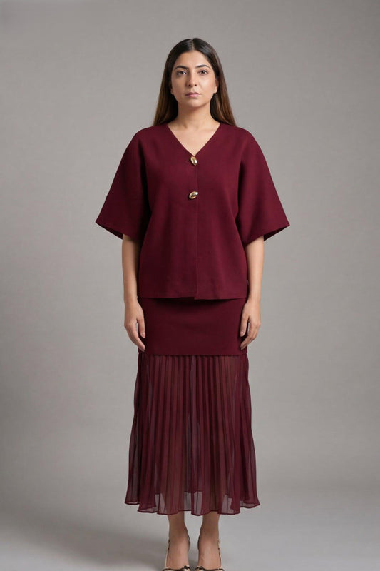 A woman modeling a matching burgundy two-piece set consisting of a short-sleeve V-neck top with a decorative front button and a pleated midi skirt, shot against a solid gray studio background.