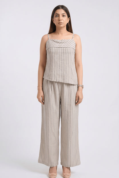 A woman modeling a matching two-piece set consisting of a sleeveless camisole top and wide-leg trousers, both in a light beige fabric with thin vertical gray stripes, shot against a plain light gray background.