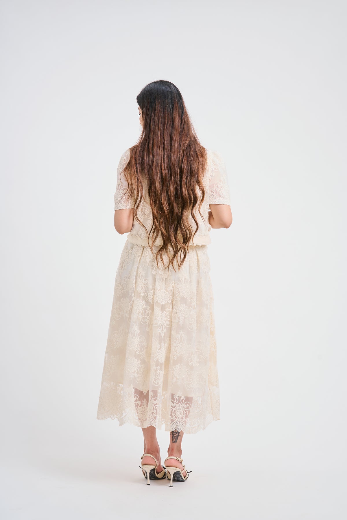 A woman viewed from behind wearing a cream lace short-sleeve midi dress and black high-heeled sandals against a plain white studio background.