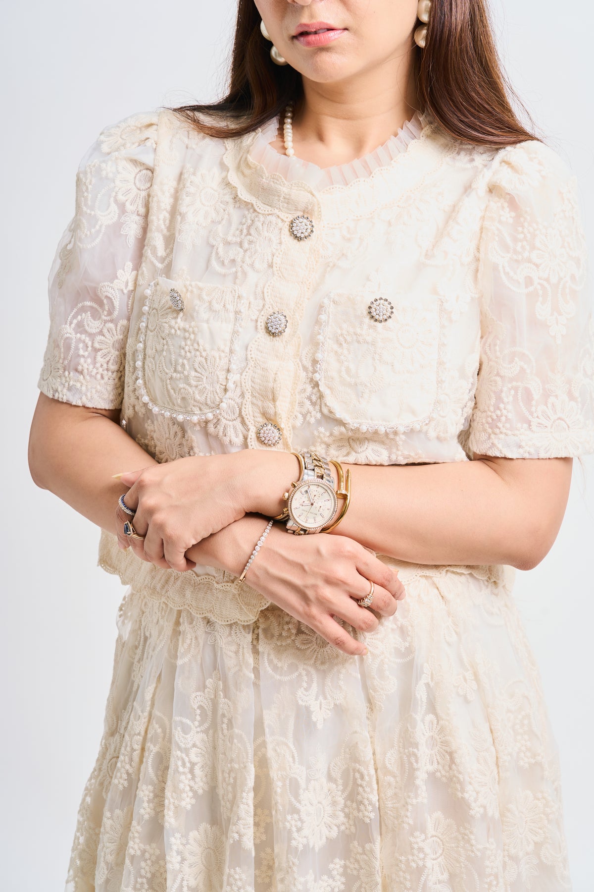 A woman wearing a cream-colored lace short-sleeve jacket with decorative buttons and a matching lace skirt, accessorized with a gold watch and rings, against a light gray background.
