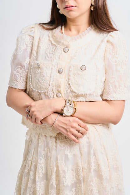 A woman wearing a cream-colored lace short-sleeve jacket with decorative buttons and a matching lace skirt, accessorized with a gold watch and rings, against a light gray background.