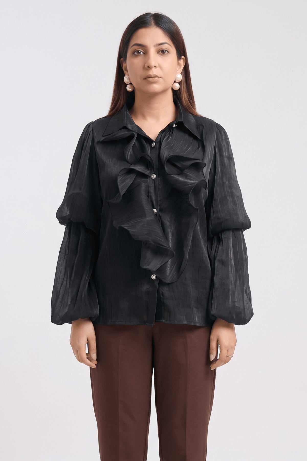 A woman modeling a black long-sleeve blouse with dramatic ruffles and balloon sleeves, paired with brown trousers, against a plain light gray studio background.