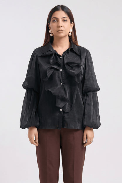 A woman modeling a black long-sleeve blouse with dramatic ruffles and balloon sleeves, paired with brown trousers, against a plain light gray studio background.