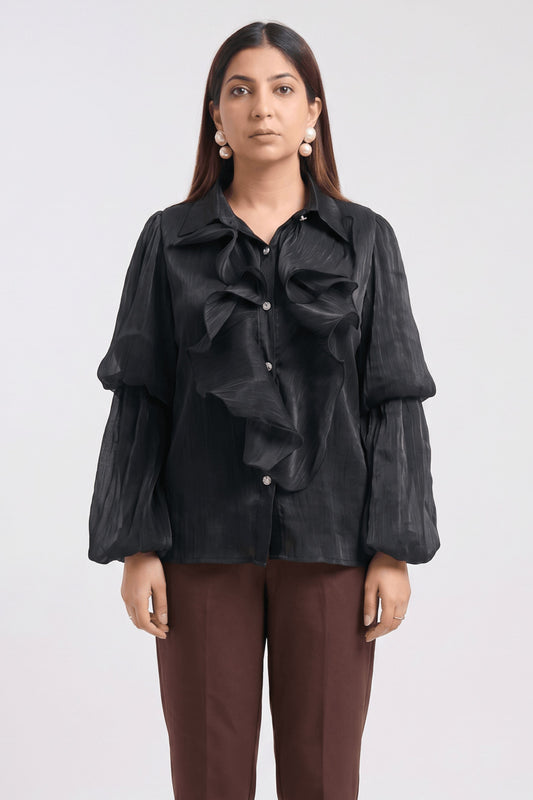 A woman modeling a black long-sleeve blouse with dramatic ruffles and balloon sleeves, paired with brown trousers, against a plain light gray studio background.