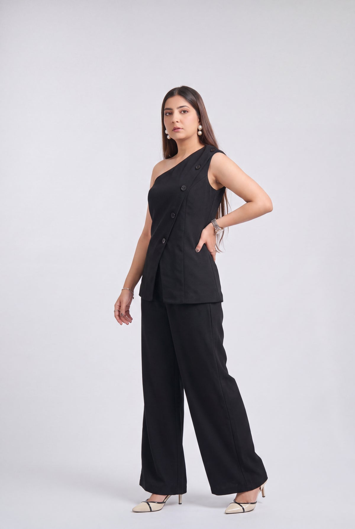 A woman modeling a black two-piece outfit consisting of a one-shoulder sleeveless top with buttons and matching wide-leg pants, photographed against a light gray studio background.