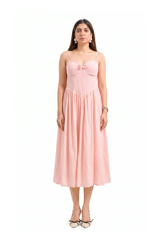 A woman modeling a light pink midi dress with spaghetti straps, a sweetheart neckline, a bow detail at the bust, and a gathered skirt, standing against a plain white background.