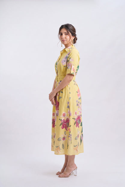 A woman modeling a yellow short-sleeve midi dress with a colorful floral print, standing against a plain light gray studio background.
